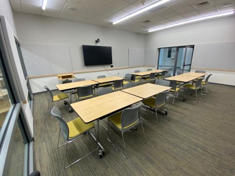Medium sized room with tables, chairs, AV screen and white board. Glass walls face out to the library.